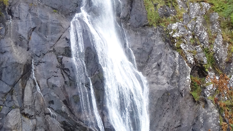 A very high and thin waterfall cascades down the dark bare rocks in the centre. On either side at the top of the falls, trees and other vegetation cling to the cliff side. At the very bottom left a person is just visible, showing the massive scale of the falls (at 120 feet tall).
