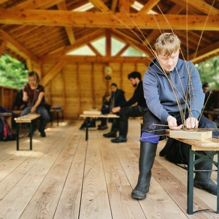 An large wooden outdoor classroom with benches dotted around with young people working with willow. Front right of the picture is James, one of the trainees. He is sat on a bench with a block of wood in front of him, and lots of willow sticking up out of it, as he winds an extra piece around his supports to build a willow bird feeder.