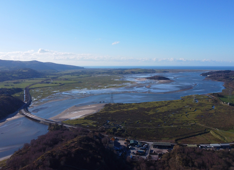 An arial photo of the estuary at Gwaith Powdwr. The river flows between open areas of green fields, with shallow patchy sand bars, and semi-flooded fields at it's borders. A road bridge crosses the river upstream, connecting both sides in a sweeping curve. Hills and wooded land enclose the estuary on one side, the sea on the other.