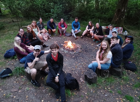 17 Mon Gwyrdd forum members sitting around the campfire in the woods smiling and looking at the camera