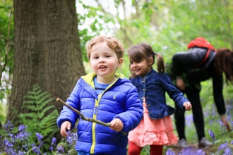 A small boy carrying a stick, walking through a woodland with large old trees and bluebells carpeting the floor. Behind him is a young girl running up to him, and a woman bending down to look at the flowers.