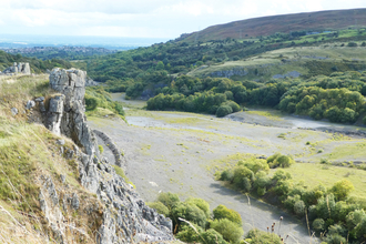 A quarry area now overgrown with vegetation, trees and other larger plants, but still with large bare patches of ground. To the left there is a steep rockface, with grasses growing everywhere. To the right hills and woodlands rise up to enclose the area. In the very far left background a town can be seen, along with fields fading into the horizon and meeting the clouds.