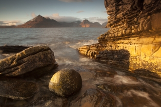 Small waves lap around large rocks at the waters edge of a beach. The sun is casting long shadows and lighting up everything with a yellow orange glow. Across the water is a large mountain range, with dark rocky appearance, except where the sunlight highlights it's faces in yellow light.