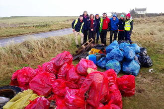 8 North Wales Wildlife Trust staff in branded clothing and high vis, smiling. They are stood behind a large pile of separated blue and red rubbish bags, and some larger debris in a clearing of dune grasses.