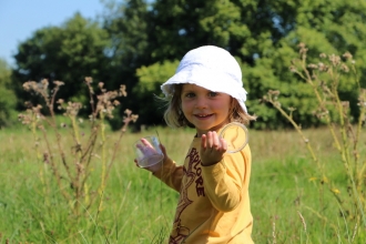 A young girl in a yellow top and white sun hat, stood in a field with tall grasses and trees in the distance, holding up a clear plastic bug pot