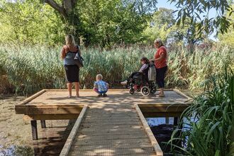 Four people, including a small child bending down for a better view, and a young girl in a wheelchair, at the end platform of the boardwalk at Big Pool Wood. Below them is one of the large ponds, with reeds and grasses further out in the water, and trees in the background.