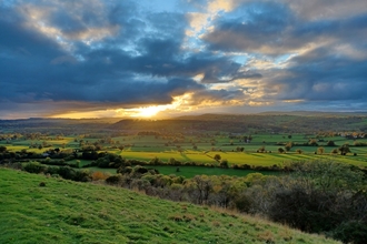 The view from Y Graig Nature Reserve across a patchwork of farm fields and distant hills. The sky is blue but clouded, with bright evening sun breaking through just above the horizon, casting long shadows and tinting the landscape with a golden glow.