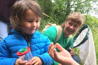 A small child holding a bug pot, looking at a grasshopper on an adults hand in front of them. Another adult holding a bug sweep net is crouched behind them looking at the child. They are at the edge of a field, with trees and long grass behind them.
