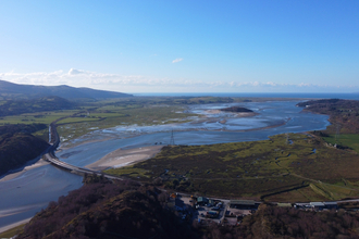 An arial photo of the estuary at Gwaith Powdwr. The river flows between open areas of green fields, with shallow patchy sand bars, and semi-flooded fields at it's borders. A road bridge crosses the river upstream, connecting both sides in a sweeping curve. Hills and wooded land enclose the estuary on one side, the sea on the other.