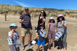 A group of 3 adults and 3 children on a sandy beach. Each is holding up an egg case or mermaids purse, The children also have buckets and ID guides. Behind them is a grassy sand dune and bright blue sky.