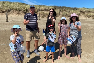 A group of 3 adults and 3 children on a sandy beach. Each is holding up an egg case or mermaids purse, The children also have buckets and ID guides. Behind them is a grassy sand dune and bright blue sky.
