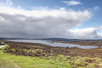 A large lake surrounded by moorland, mainly brown with dried up heather, but some green grasses in the foreground and fields to the left. A small track and fence line separates the fields from the reserve. On the far side of the lake hills rise to enclose the area, with a shadow cast over them by immense fluffy clouds. Above them the sky is a deep blue, and the sun is illuminating the cloud edge so it glows bright white.