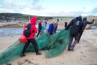 A group of 7 people hauling a washed up fishing net off a beach. In the background many more people can be seen litter picking during a very large beach clean at plastoff 2022