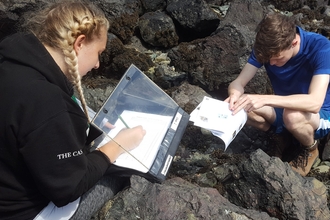 Two trainees sat on a black rocky beach covered in seaweed taking notes
