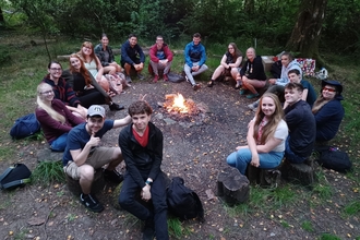 17 Mon Gwyrdd forum members sitting around the campfire in the woods smiling and looking at the camera