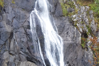 A very high and thin waterfall cascades down the dark bare rocks in the centre. On either side at the top of the falls, trees and other vegetation cling to the cliff side. At the very bottom left a person is just visible, showing the massive scale of the falls (at 120 feet tall).