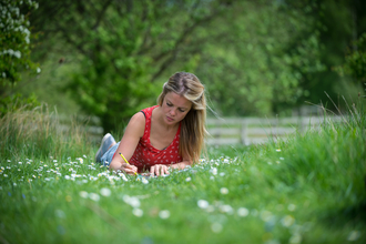 A woman in a red top with long blonde hair, lying on her front, propped up on her elbows and writing in a note book on the ground. She is in a field, with lots of grass and daisies, behind her there is a fence and trees that are blurry.