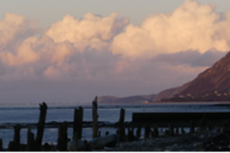 A view of the estuary from the Spinnies Aberogwen reserve. On the ground in the front our several wooden posts and to the right you can see the side of a mountain cliff up ahead. The clouds are hanging low bathed in pinks and yellow as the sun is setting. 