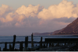 A view of the estuary from the Spinnies Aberogwen reserve. On the ground in the front our several wooden posts and to the right you can see the side of a mountain cliff up ahead. The clouds are hanging low bathed in pinks and yellow as the sun is setting.