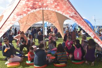 A group of people including many children sat under an orange, 5 legged, starfish tent. Some are on chairs, some on cushions on the floor, all listening to one woman on a wooden chair telling a story. In the background are more tents and gazebos with people wandering around on a grassy area.