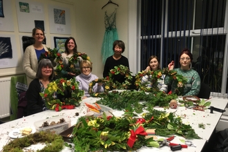 A group of ladies holding up hand made Christmas wreaths, in front of them is a table full of greenery, ribbons and tools for making wreaths.