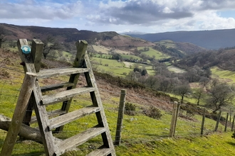 A wooden stile with a footpath marker crosses a fence between 2 fields. On the other side is a valley of patchwork fields and trees lit in bright sunlight. Hills raise up on either side cast in shadow by the fluffy white clouds above.