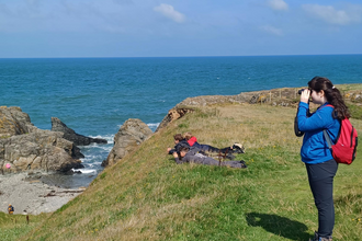 A girl stood on a grassy cliff facing left with binoculars looking down into a cove. Behind her, 3 boys are lying down near the cliff slope on their fronts to get a better view. A small cove with stony beach is visible to the left below them, and a wide blue sea behind them.