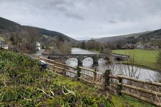 A river with a small stone arch bridge crossing it, set in a valley with farm fields and sparse houses. The hills in the distance seem to encircle the whole area.