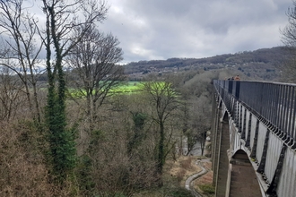 A large aqueduct with stone columns and metalwork bridge façade. It crosses a valley with a river, and shallow sloping sides covered in dark trees with no leaves.