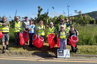 A group of 10 people smiling at the camera on a road verge. They are wearing high vis, holding up litter pickers and red bin bags. On has a sign with the North Wales Wildlife Trust logo on it.