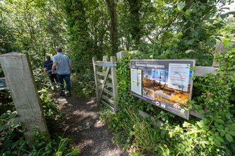 A path through an open wooden gate, with 2 people walking away into a woodland. Next to the gate is a large sign which reads 'Spinnies Aberogwen', with pictures of the reserve, Egret, and Kingfisher.