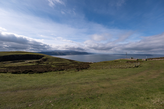 A view from the Great Orme, Grass and heathland hills drop off down to the sea. In the distance the mainland curves around enclosing the ocean like a bay. Above clouds are gathered in rows, fluffy and dark, with a deep blue sky.