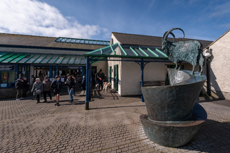 A large building with metal and glass roof verandas, brick paving outside, and a metal goat sculpture. There is a group of people and dogs leaving the building on a clear sunny day.