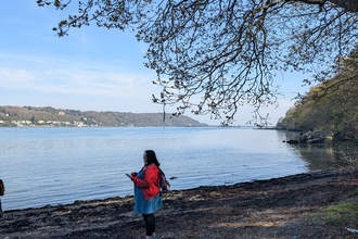 Woman wearing red top standing under a tree on the shore of the Menai Strait. Blue sky.