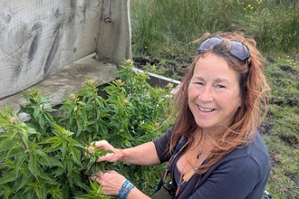 Jules Cooper, a lady with reddish brown hair and lightly tanned white skin, wearing blue jeans and a charcoal grey top. She smiles at the camera while kneeling down and handling a plant growing from the back of a wooden bench in a field.