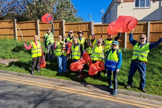 11 volunteers in high vis vests stood on a grass verge at the side of a road. They are holding up litter pickers and red bin bags cheering