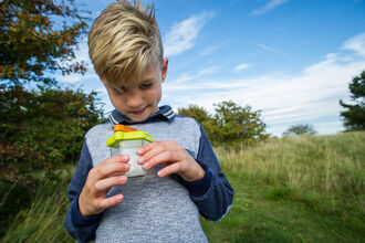 A young boy wearing grey and blue, stood in a field holding a clear plastic bug pot, looking into the yellow and orange magnification lid.