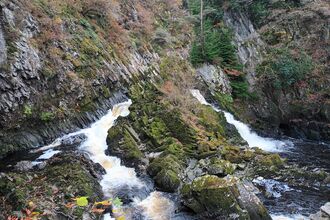 Sheer rocky river banks meet in a series of small waterfalls and rapids. The river splits in 3 directions around large rocky outcrops, with vegetation, evergreen and bare branched trees of winter clinging to the slopes.