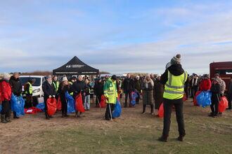 A large gathering of people in winter clothing, in a sandy dune edge car park. Most are holding litter pickers and blue or red bags, one at the front is wearing high visibility yellow and addressing the crowd through a megaphone. The day is clear and cold with a pale blue sky and light streaky clouds.