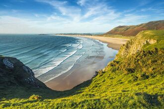 A wide coastal landscape viewed from a grassy cliff, overlooking a long sandy beach with gentle waves rolling in from the blue ocean. Rolling green hills and rocky cliffs frame the shoreline under a partly cloudy sky.
