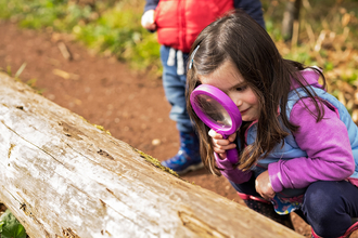 A young girl wearing pink and purple, crouching down to look through a large pink magnifying glass at a big log with moss on it. A second child is out of focus stood behind her.