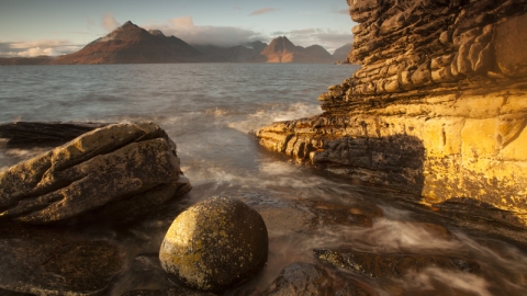 Small waves lap around large rocks at the waters edge of a beach. The sun is casting long shadows and lighting up everything with a yellow orange glow. Across the water is a large mountain range, with dark rocky appearance, except where the sunlight highlights it's faces in yellow light.