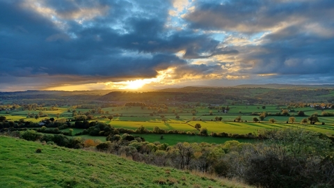 The view from Y Graig Nature Reserve across a patchwork of farm fields and distant hills. The sky is blue but clouded, with bright evening sun breaking through just above the horizon, casting long shadows and tinting the landscape with a golden glow.
