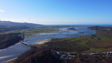 An arial photo of the estuary at Gwaith Powdwr. The river flows between open areas of green fields, with shallow patchy sand bars, and semi-flooded fields at it's borders. A road bridge crosses the river upstream, connecting both sides in a sweeping curve. Hills and wooded land enclose the estuary on one side, the sea on the other.
