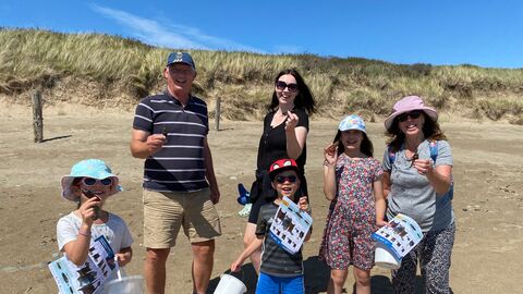 A group of 3 adults and 3 children on a sandy beach. Each is holding up an egg case or mermaids purse, The children also have buckets and ID guides. Behind them is a grassy sand dune and bright blue sky.