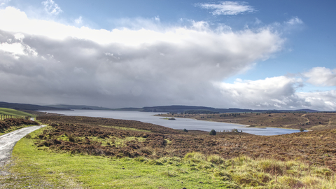 A large lake surrounded by moorland, mainly brown with dried up heather, but some green grasses in the foreground and fields to the left. A small track and fence line separates the fields from the reserve. On the far side of the lake hills rise to enclose the area, with a shadow cast over them by immense fluffy clouds. Above them the sky is a deep blue, and the sun is illuminating the cloud edge so it glows bright white.