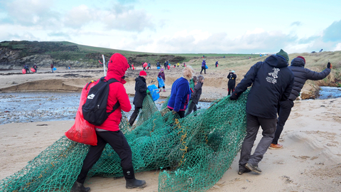 A group of 7 people hauling a washed up fishing net off a beach. In the background many more people can be seen litter picking during a very large beach clean at plastoff 2022