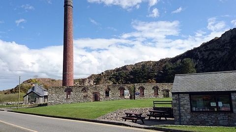 At the bottom right of picture is a small building, the NWWT shop) and patio area with picnic benches. Infront of them a road runs from right to left. The centre of the picture features a very old wall with lots of barred windows, and slightly left reaching all the way to the top of the picture is a large old style industrial chimney. In the background the rocky cliffs that surround the park rise in height from left to right. The sky above them is a bright blue with lots of fluffy white clouds.