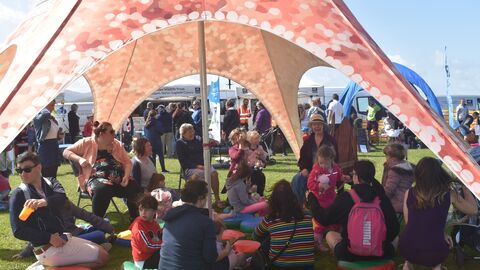 A group of people including many children sat under an orange, 5 legged, starfish tent. Some are on chairs, some on cushions on the floor, all listening to one woman on a wooden chair telling a story. In the background are more tents and gazebos with people wandering around on a grassy area.