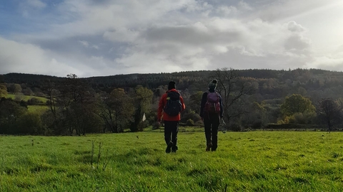 2 people in outdoor clothing, woolly hats and waterproofs, walking through an open field towards a hilly landscape with lots of tree cover.